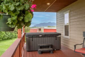 A covered wooden deck at 868 Middle Road features a hot tub and small steps, overlooking green grass and distant mountains. A hanging plant and red building are visible, with a single chair by the house’s beige siding.