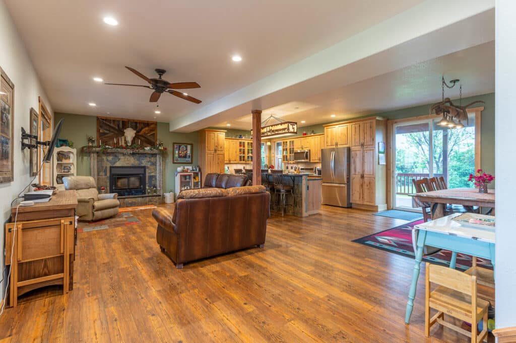 Open-plan living room and kitchen at 868 Middle Road with wood floors, leather sofa, fireplace, ceiling fan, and dining area. Natural light streams in through large windows and doors leading to a deck with views of greenery outside.