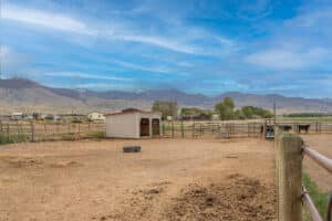A fenced dirt paddock at 868 Middle Road features a small shelter, trough, and a few horses, set against distant mountains beneath a partly cloudy blue sky. Rural buildings and green trees complete the peaceful backdrop.