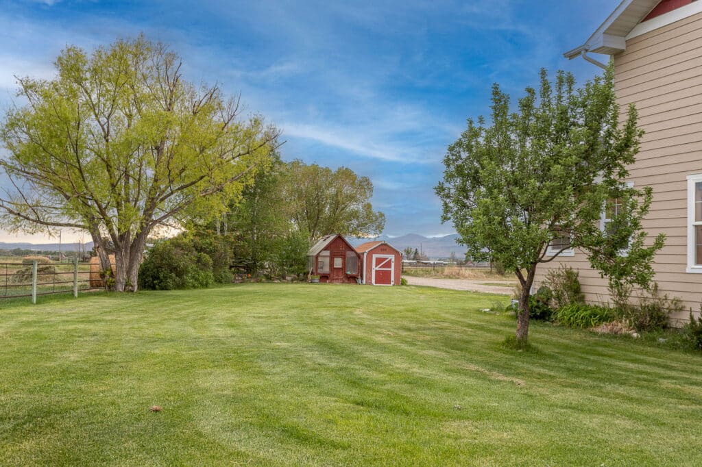 A spacious green lawn at 868 Middle Road features a large tree, a small red barn, and a beige house on the right under a blue sky with scattered clouds. A wooden fence and distant fields complete the peaceful scene.