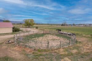 A round wooden corral with patchy grass stands in a rural area at 868 Middle Road, surrounded by open fields, a few buildings, and distant mountains under a blue sky with scattered clouds.