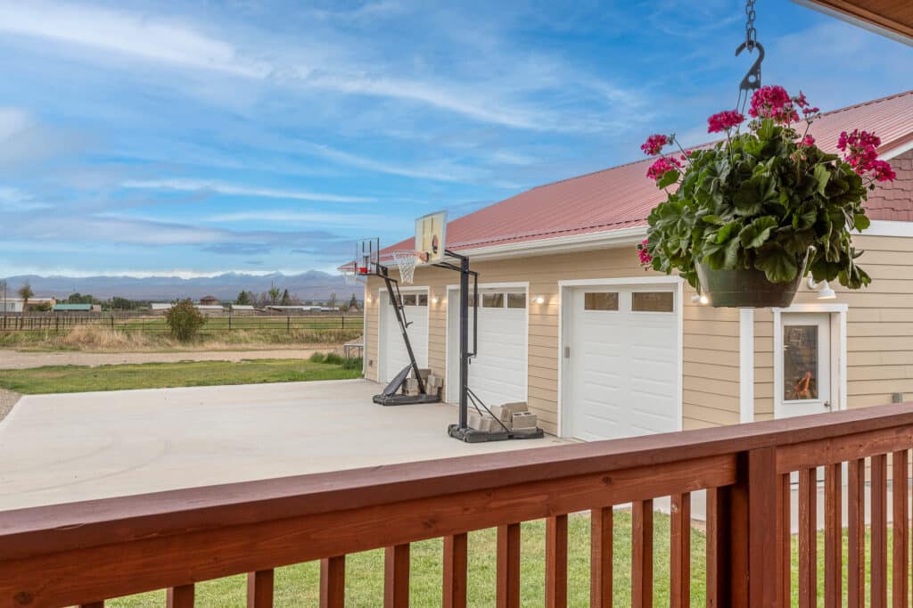 A view from a wooden deck at 868 Middle Road overlooks a concrete driveway with two basketball hoops, a beige house with a red roof, hanging flowers, and a scenic landscape in the background.