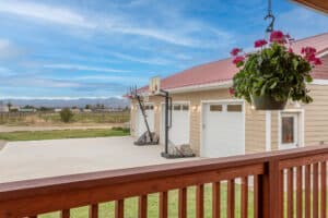 A view from a wooden deck at 868 Middle Road overlooks a concrete driveway with two basketball hoops, a beige house with a red roof, hanging flowers, and a scenic landscape in the background.