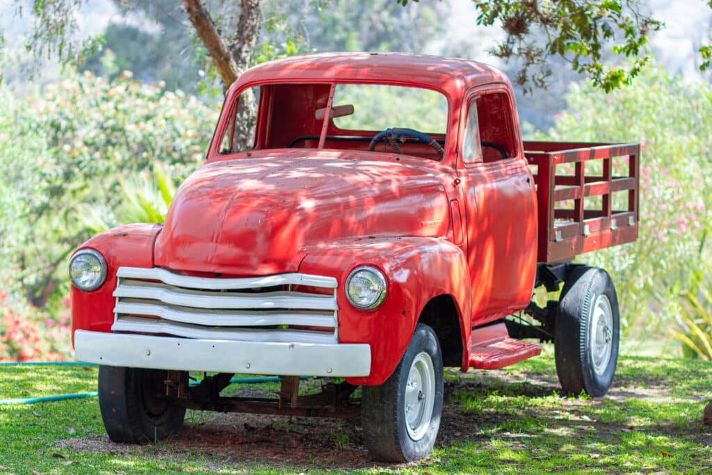 A bright red vintage pickup truck with white accents is parked on green grass under a tree, surrounded by lush greenery and sunlight filtering through the leaves. This classic vehicle stands ready to haul farm tools and essential ranch equipment, perfectly at home in its idyllic countryside setting.