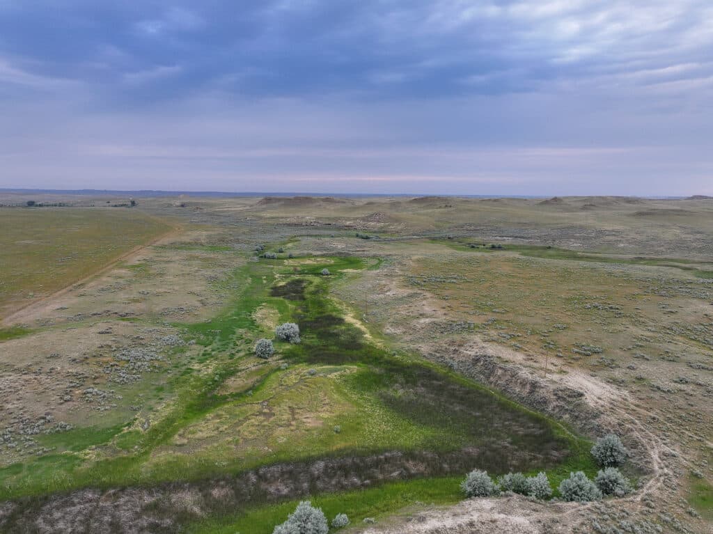 Aerial view of Antelope Springs Ranch reveals a vast, open landscape with green patches, sparse vegetation, and a cloudy sky. The terrain is mostly flat with gentle slopes and scattered shrubs.