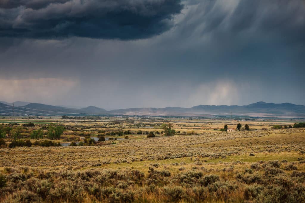 A vast open field with dry grass and sparse shrubs stretches under a cloudy, dark sky with distant rain showers; mountains and patches of green farmland are visible on the horizon, evoking the rugged beauty surrounding Corps of Discovery Ranch.