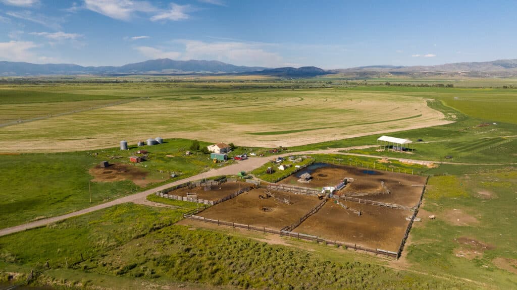 Aerial view of Discovery Ranch, a rural farm with fenced animal pens, a house, several outbuildings, and open green fields under a blue sky with distant mountains.