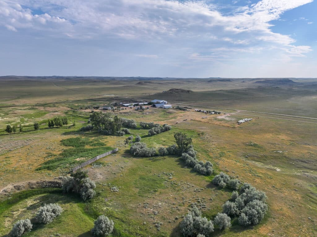 Aerial view of a rural landscape with scattered trees, green fields, and the wide, open sky with light clouds, featuring the distant cluster of buildings at Antelope Springs Ranch.