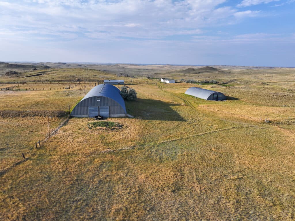 Aerial view of a rural landscape with two large, curved-roof metal barns, grassy fields, fences, and a small white building in the distance under a blue sky with clouds at Antelope Springs Ranch.