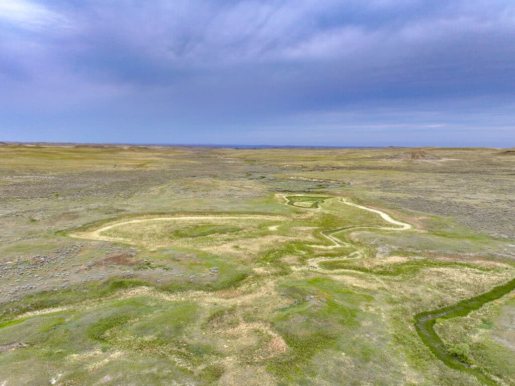 Aerial view of a winding creek cutting through the vast, flat grassland of Antelope Springs Ranch under a cloudy blue sky, with patches of green and brown vegetation visible across the landscape.