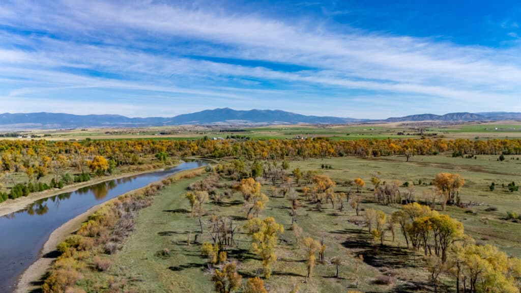 An aerial view of a winding river flowing through the golden autumn landscape of Corps of Discovery Ranch, with distant mountains under a blue sky.