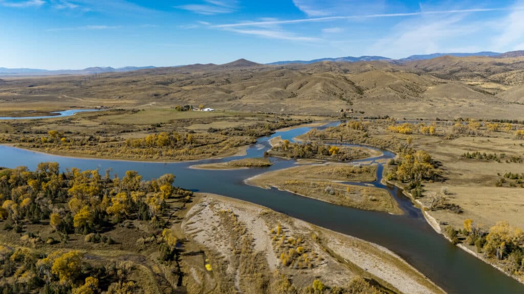An aerial view of a winding river flowing through the Corps of Discovery Ranch, surrounded by a dry, hilly landscape with patches of yellow autumn trees.