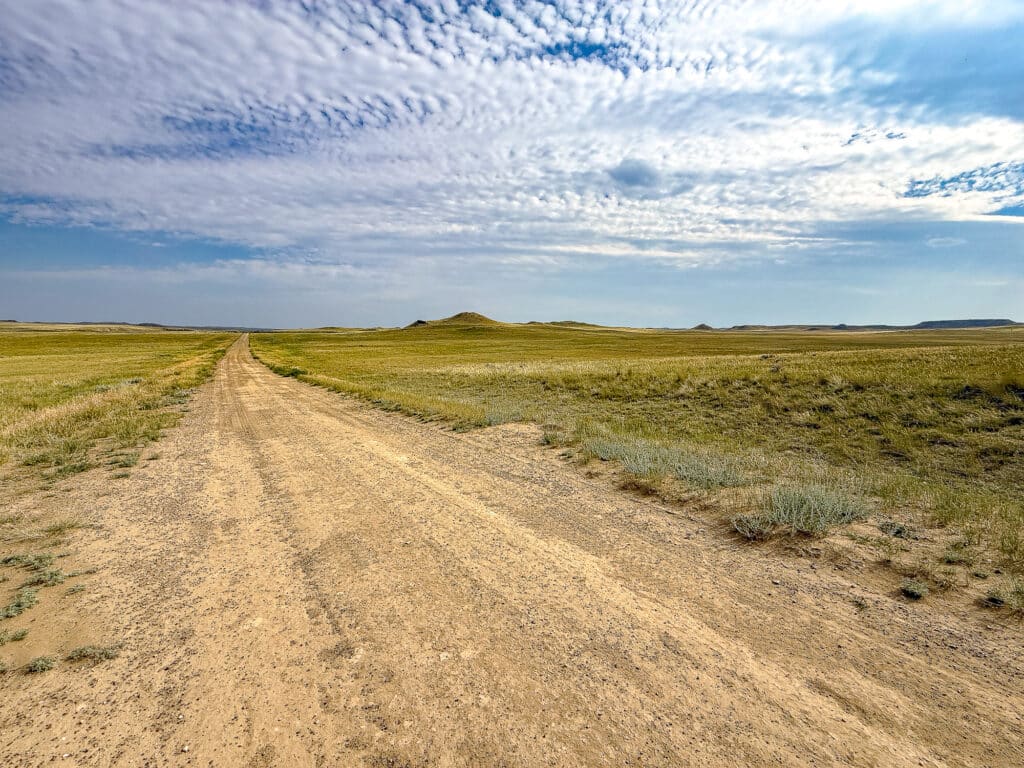A dirt road stretches through a vast, flat grassy plain under a blue sky with scattered clouds, with low hills visible in the distance—a tranquil scene typical of Antelope Springs Ranch.