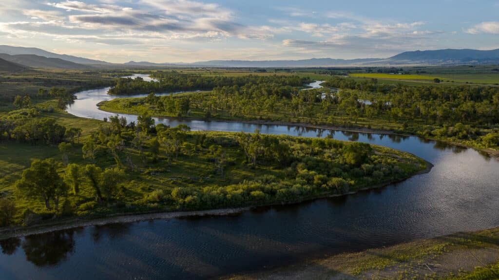 A winding river flows through a lush green landscape with trees and fields, mountains in the distance, and a partly cloudy sky at sunset, evoking the tranquil beauty of Discovery Ranch.