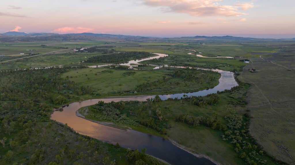 A winding river flows through a green landscape under a colorful sunset sky, with distant mountains and scattered clouds in the background, evoking the tranquil beauty of Corps of Discovery Ranch.