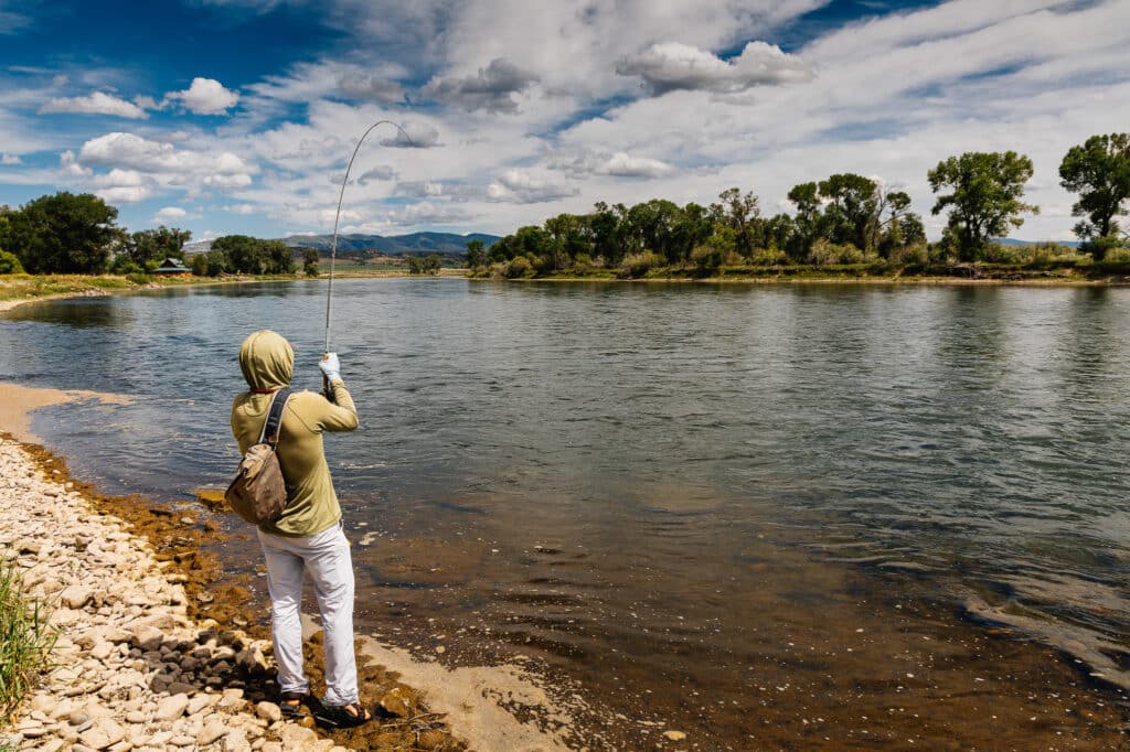 A person in light clothing and a hood fishes by a wide, calm river with a rocky shore at Corps of Discovery Ranch, surrounded by green trees and mountains under a partly cloudy blue sky.