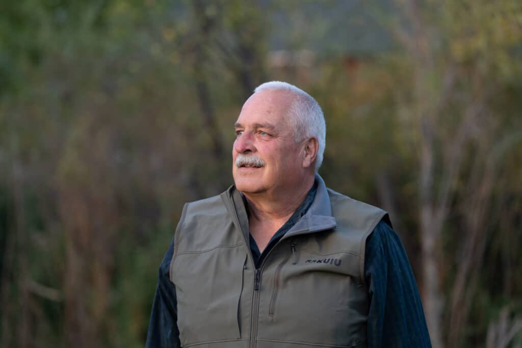An older man with short gray hair and a mustache wears a gray vest and dark shirt, standing outdoors with blurred trees and greenery in the background. This is Jack McInerney of Janicki Industry, thoughtfully surveying the site of his recent land purchase.