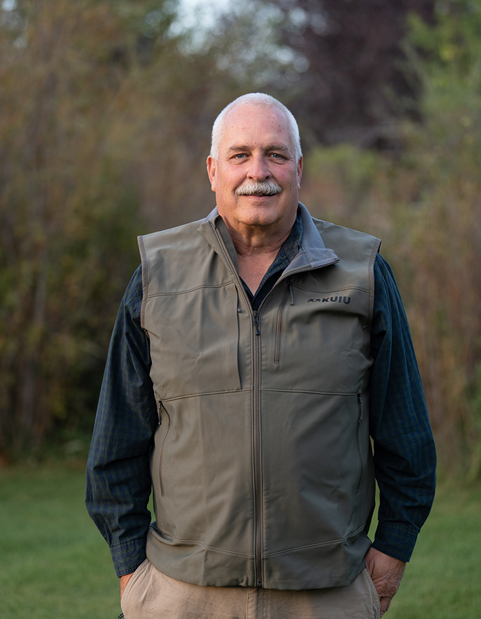 Older man with a mustache wearing a gray vest and dark shirt stands outdoors on grass, with trees and bushes blurred in the background, reminiscent of land purchase discussions near Great Falls for Janicki Industry.