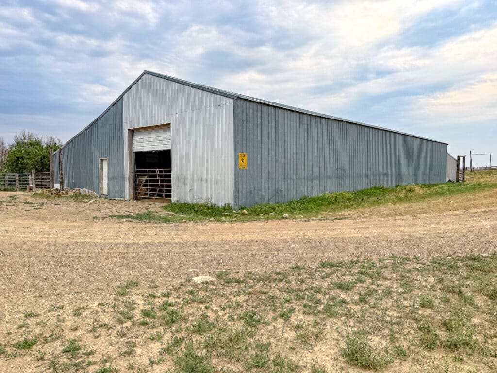 A large gray metal barn with a partially open garage door sits beside a dirt road under a cloudy sky, with grass and open fields surrounding the building at Antelope Springs Ranch.