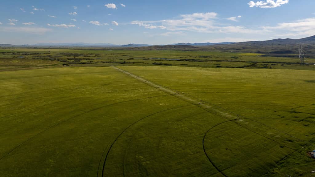 A vast green field with faint circular crop marks and a straight dirt path, set against distant hills under a blue sky with scattered clouds, evokes the open landscapes reminiscent of Discovery Ranch explored by the Corps of Discovery.