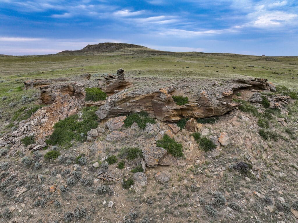 A rocky outcrop with patches of green vegetation on a grassy plain under a blue sky with scattered clouds; beyond, the landscape of Antelope Springs Ranch unfolds as a low hill rises in the distance.