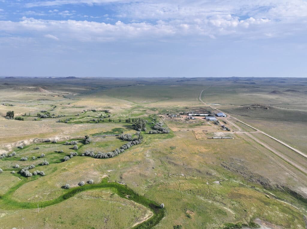 Aerial view of Antelope Springs Ranch, a remote property surrounded by vast open plains, scattered trees, dirt roads, and distant hills under a partly cloudy sky.