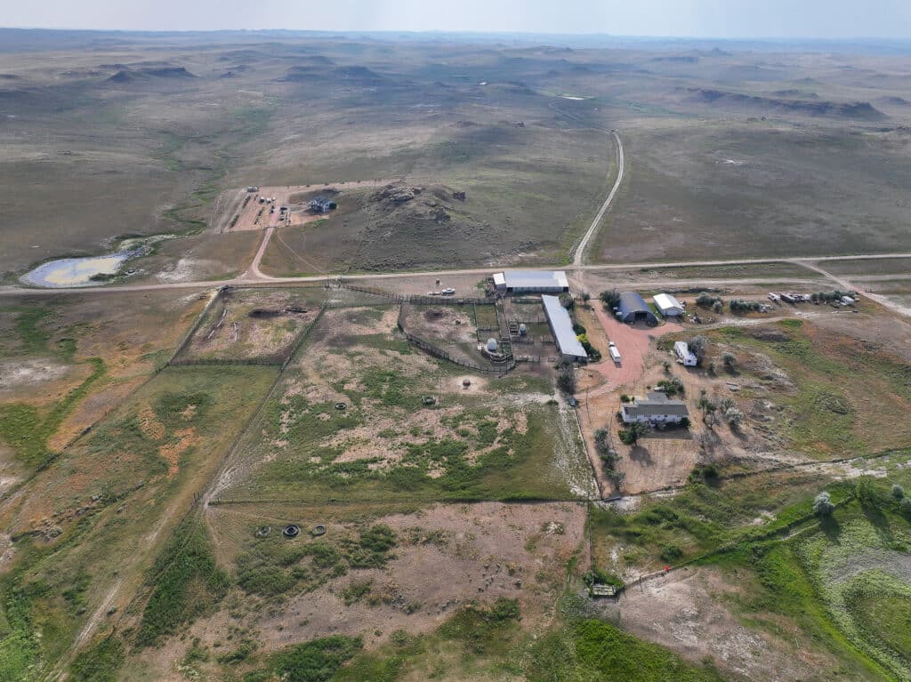 Aerial view of Antelope Springs Ranch with several buildings, dirt roads, fenced areas, and open fields, set in a vast, dry, and lightly rolling landscape under a hazy sky.