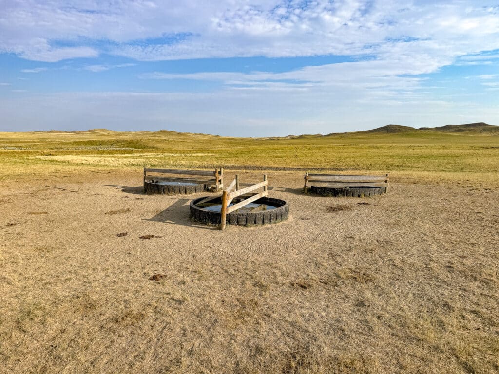 A circular livestock water trough made from large tires stands in a dry, grassy field under a partly cloudy sky at Antelope Springs Ranch, surrounded by open plains and distant hills.