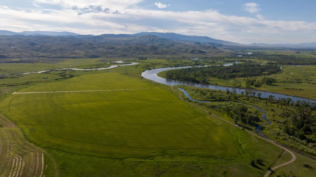 A winding river flows through wide green fields and patches of trees, with hills and distant mountains under a partly cloudy sky, evoking the timeless beauty of a ranch landscape reminiscent of the Corps of Discovery’s storied journeys.