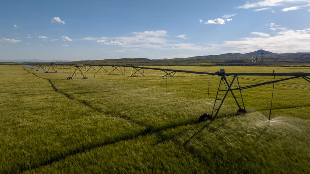 A large irrigation system waters a green field at the Corps of Discovery Ranch, set under a blue sky with scattered clouds and hills in the background.