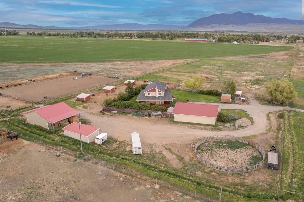 Aerial view of 868 Middle Road, a rural farm with a house, several red-roofed barns, a round pen, fenced areas for animals, and green fields nestled beneath mountains and a blue sky.