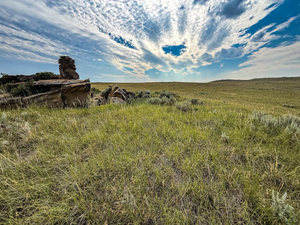 A grassy plain stretches to the horizon under a dramatic sky with scattered clouds, and a rock formation stands to the left side of the image, capturing the tranquil beauty of Antelope Springs Ranch.
