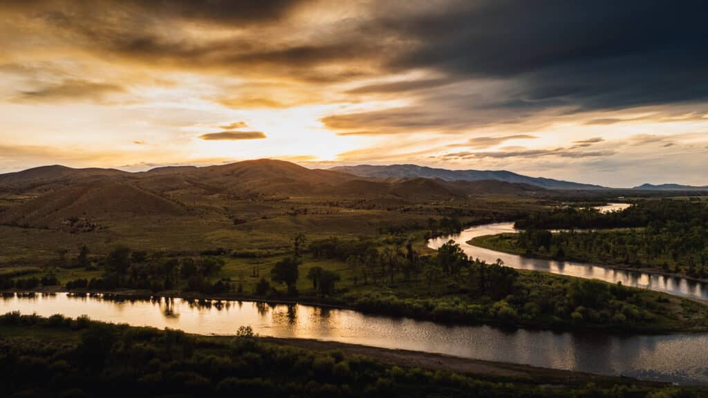 A winding river flows through the green landscape of Corps of Discovery Ranch at sunset, with dramatic clouds in the sky and hills in the background.