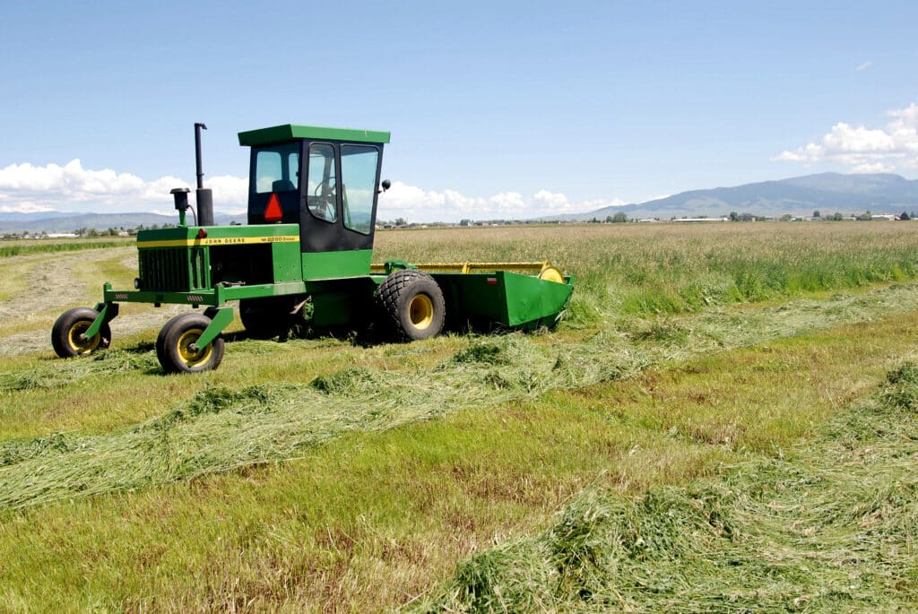 A green farm tractor with a cab, one of the most essential pieces of ranch machinery, cuts grass in a large, open field under a blue sky with scattered clouds and mountains in the background.