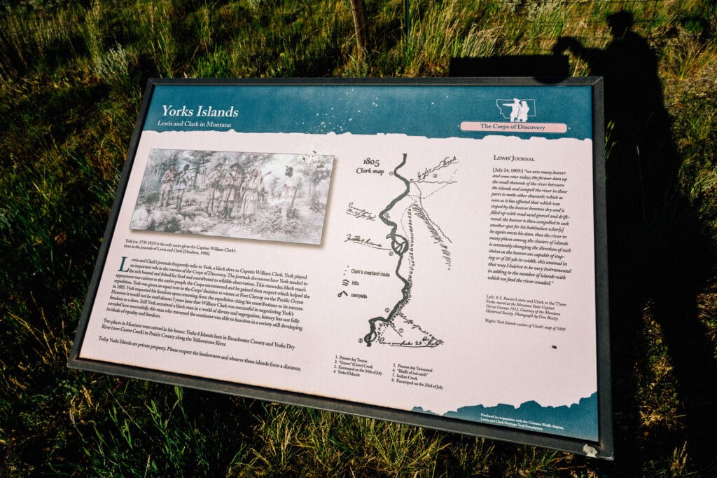 Interpretive sign titled Yorks Islands with a historical sketch, a map, and text describing the Corps of Discovery during the Lewis and Clark expedition. The sign is outdoors on grass, with a person's shadow visible on the right.
