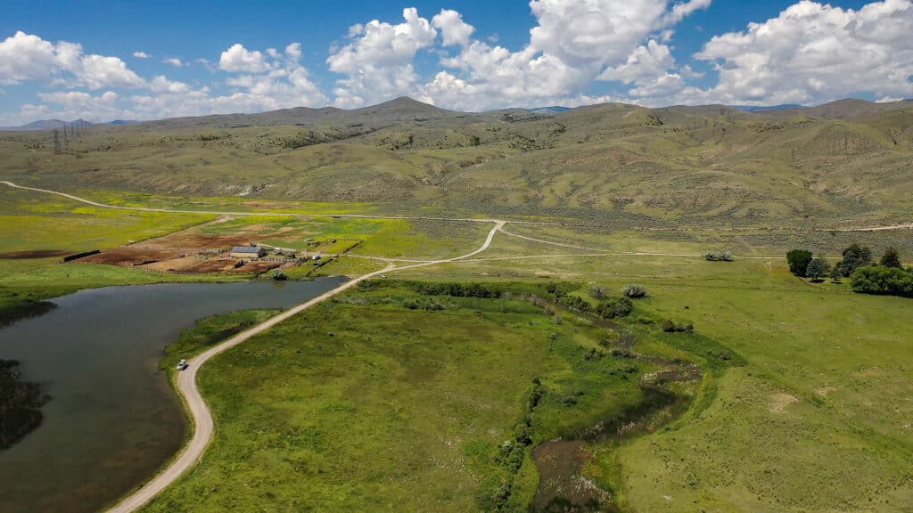 Aerial view of a rural landscape with green fields, a pond, a dirt road, scattered buildings, and rolling hills under a blue sky with white clouds—an idyllic scene reminiscent of the Corps of Discovery Ranch.