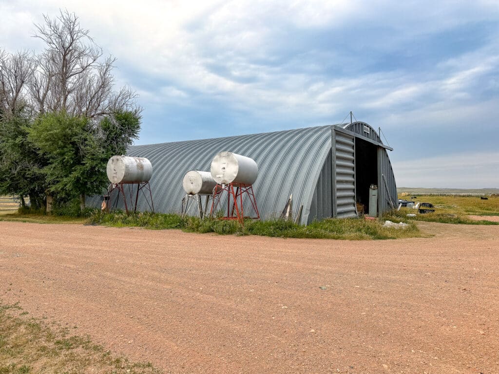 A large corrugated metal shed with three cylindrical fuel tanks on stands sits beside a dirt road at Antelope Springs Ranch, surrounded by grass and a few trees under a cloudy sky.