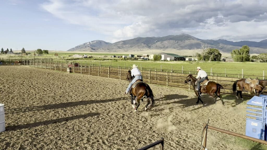 Two people on horseback ride through a dirt arena at Double T Estate with mountains in the background.