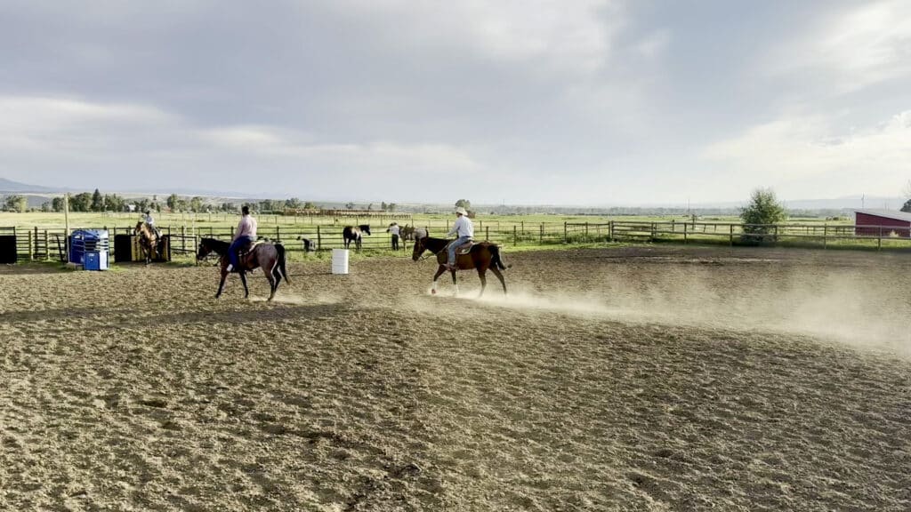 Two people riding horses in the Double T Estate outdoor arena, with other horses visible in the background.