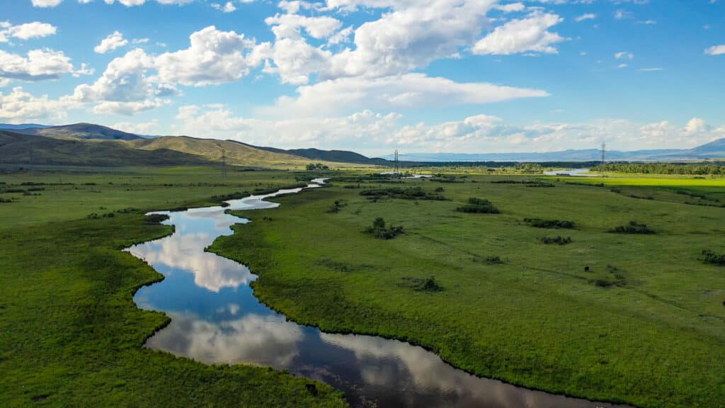 A winding river flows through a green landscape under a blue sky with scattered clouds, reflecting in the water. Rolling hills and distant electric poles are visible on the horizon, hinting at the serene expanses of Corps of Discovery Ranch.
