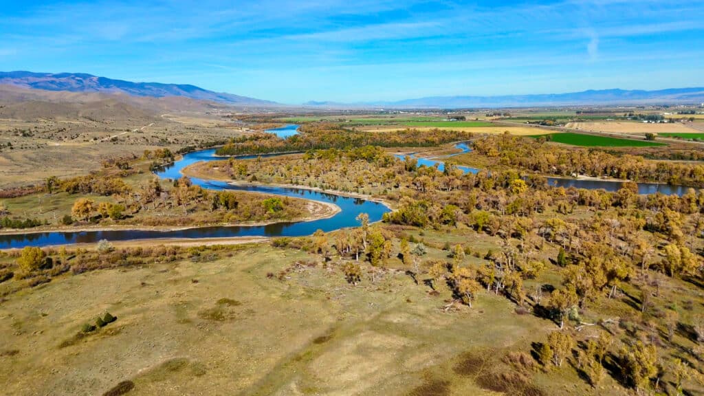 Aerial view of a winding river flowing through the dry, autumn-colored landscape of the Corps of Discovery Ranch, with mountains in the distance.