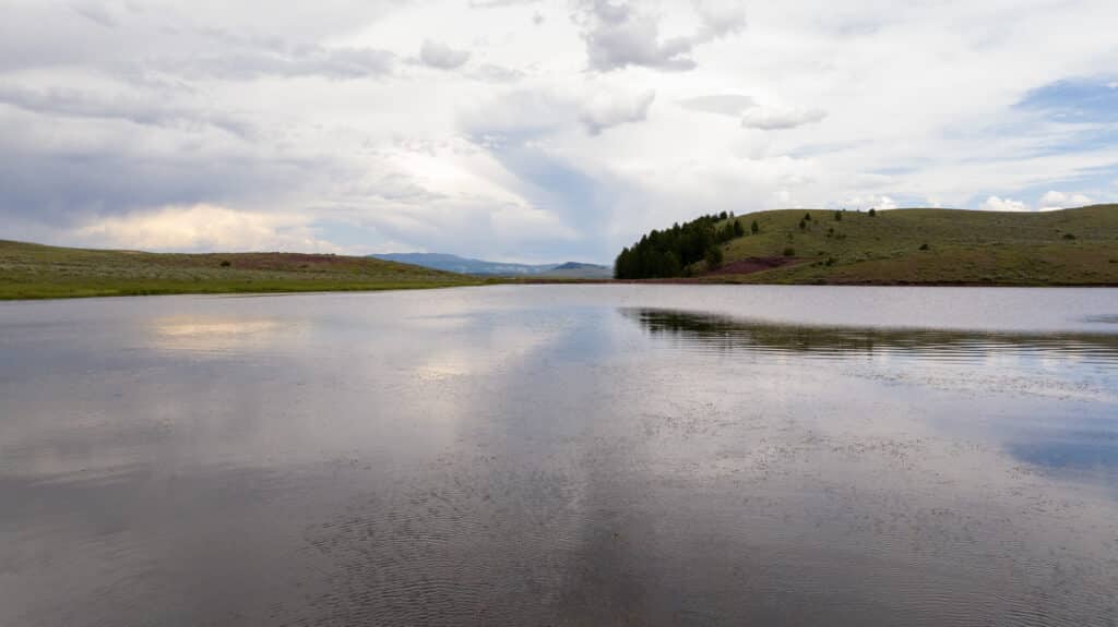 A calm lake reflects a cloudy sky, with rolling grassy hills and a cluster of trees in the distance under soft, diffused light—capturing the tranquil beauty characteristic of Battle Mountain Ranch.
