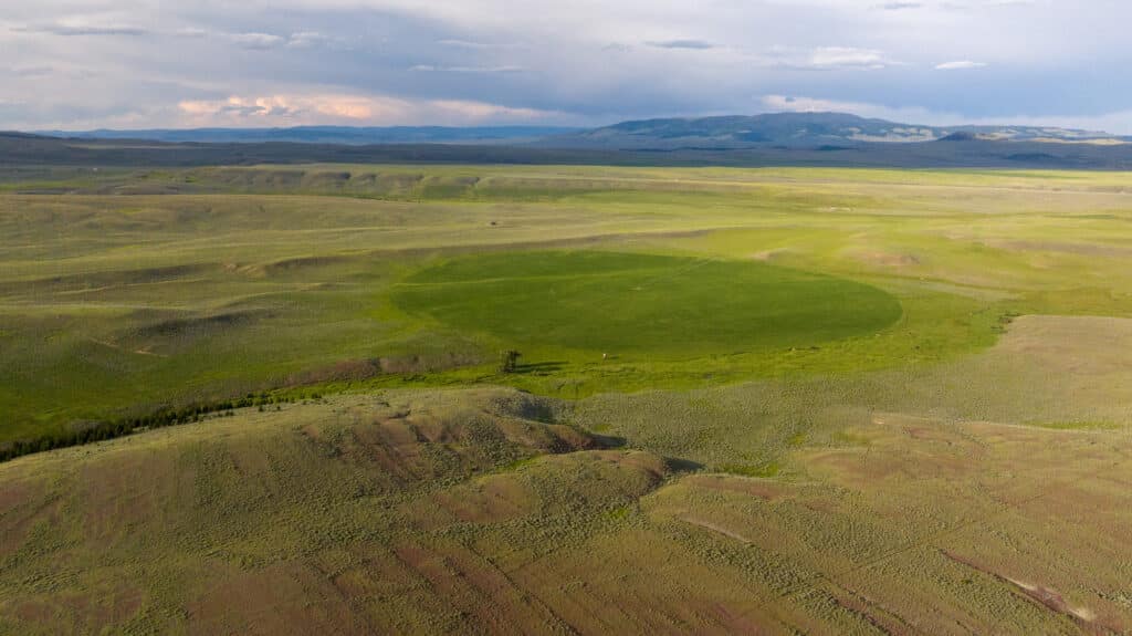 Aerial view of a vast landscape with a large, circular green field in the center—part of Battle Mountain Ranch—surrounded by dry, brown land and distant mountains under a cloudy sky.