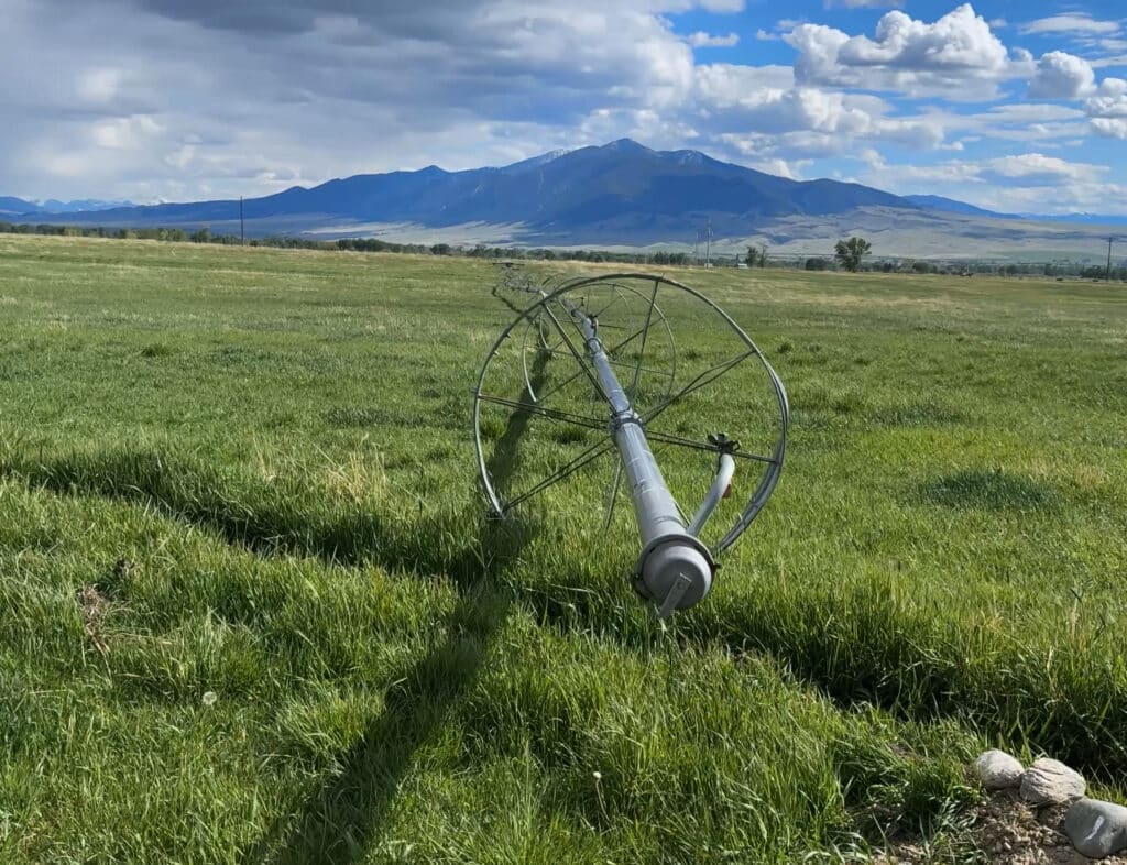 A large metal irrigation pivot lies toppled in a green field on a Horse Ranch under a partly cloudy sky, with mountains and the tranquil waters of Wisconsin Creek visible in the background.
