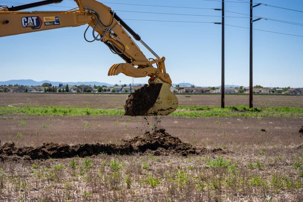 A yellow excavator digs into a field on the Janicki Industries Manufacturing Campus in Great Falls, Montana, lifting a bucket of soil with houses and mountains visible in the background under a clear blue sky.