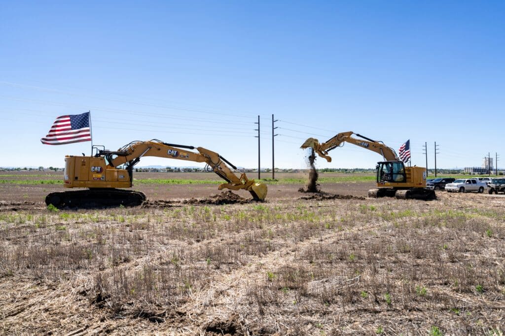 Two excavators with American flags dig into a field under a clear blue sky at the future site of the Janicki Industries Manufacturing Campus in Great Falls, Montana. Dirt is being moved, and utility vehicles are parked in the background near power lines.