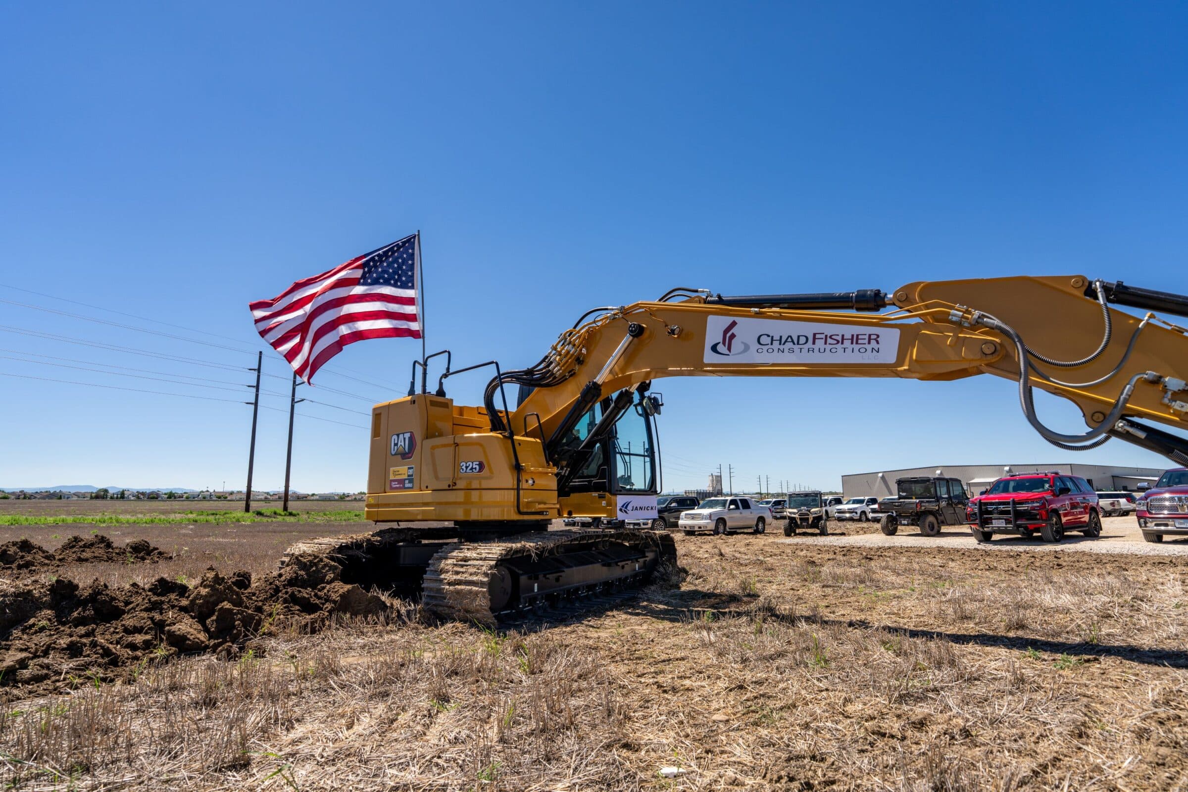 A yellow excavator with a U.S. flag attached stands on a dirt lot under a clear blue sky at the Manufacturing Campus in Great Falls, Montana. Several vehicles are parked in the background near Janicki Industries, and the excavator bears the Chad Fisher Construction logo.