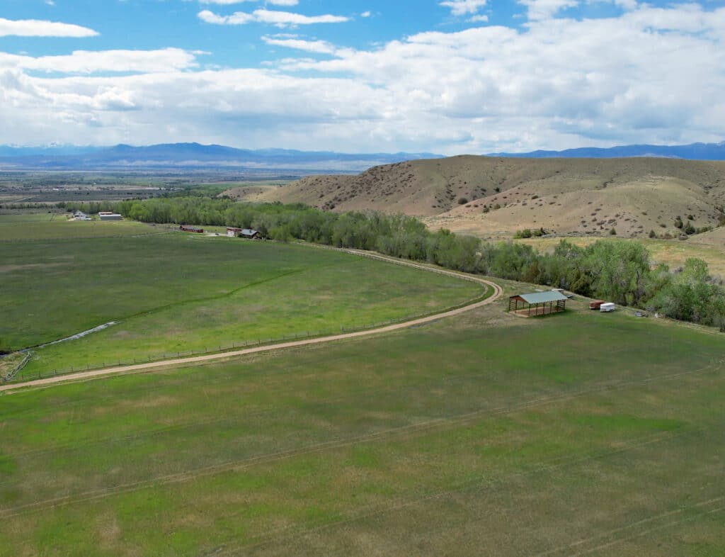 Aerial view of a large green field with a winding dirt road, a small shed, trees by the edge, rolling hills, and distant mountains under a partly cloudy sky—an idyllic scene reminiscent of a classic Wisconsin Creek horse ranch.