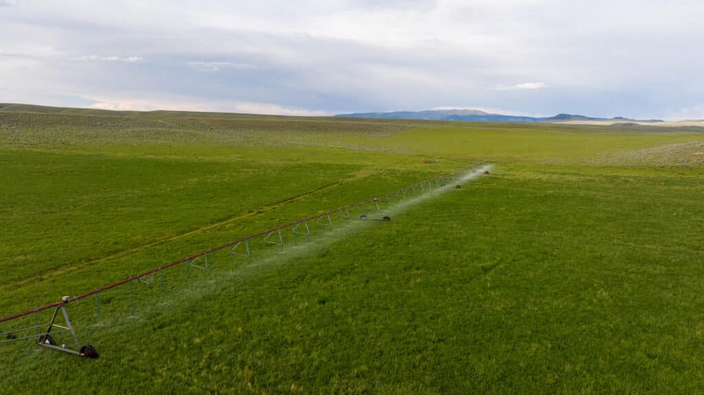 Aerial view of a green field with an irrigation system spraying water, stretching across the grass under a partly cloudy sky with distant hills in the background at Battle Mountain Ranch.