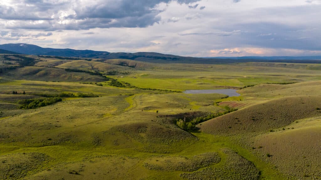 Rolling green hills with scattered shrubs and a small pond under a cloudy sky, mountains in the distant background evoke the serene beauty of Battle Mountain Ranch.
